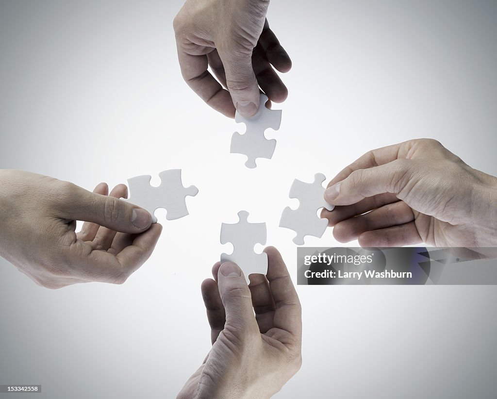 Four hands holding jigsaw puzzle pieces, directly above