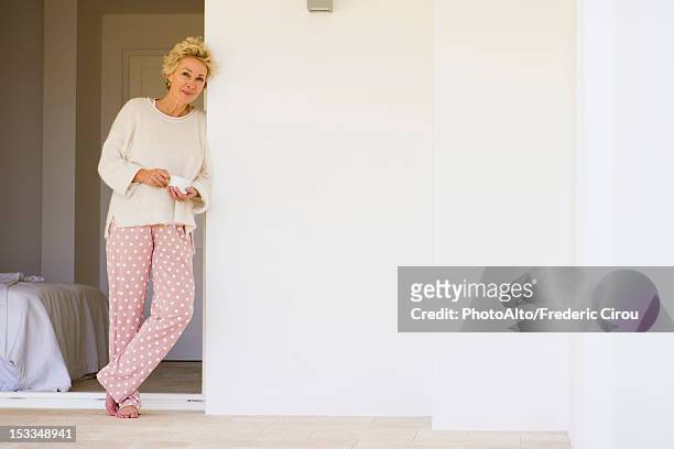 mature woman leaning against wall in pajamas with cup of coffee - pyjamas stock pictures, royalty-free photos & images