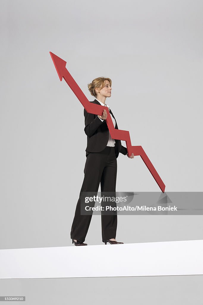 Businesswoman walking on balance beam, carrying large arrow indicating profits