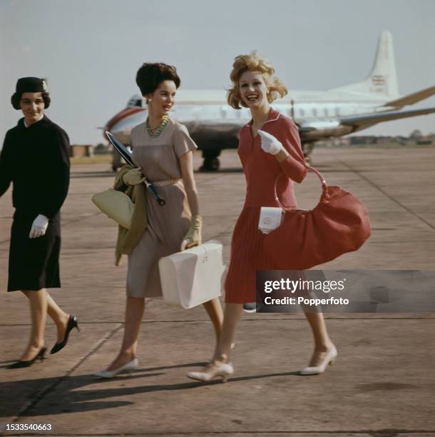 Two female fashion models walk with an air stewardess towards their aircraft at an airport, they wear, from left, a Tricel dress in coffee cream and...