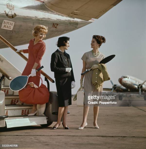 Two female fashion models posed with an air stewardess at the rear steps of an aircraft at an airport, they wear, from left, a red jersey wool top...