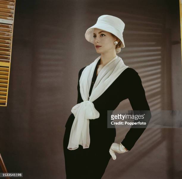 Posed studio portrait of a female fashion model wearing a fitted black skirt suit with white gloves, white cloche hat and matching white chiffon...