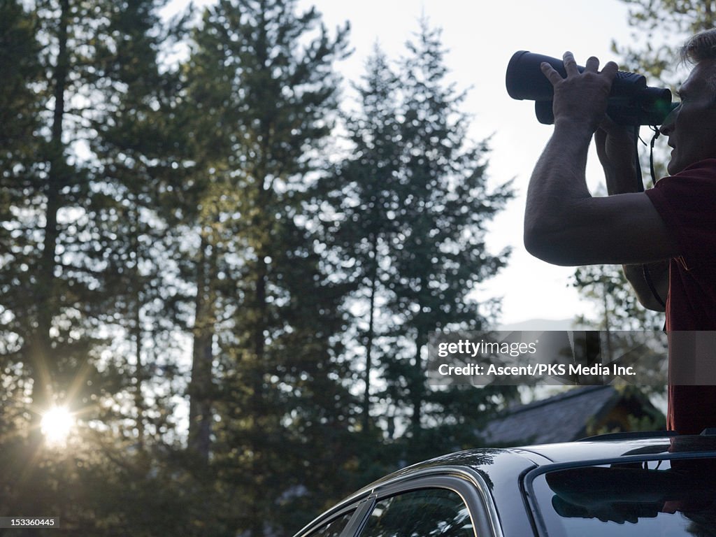 Man stands through car's skyroof, uses binoculars