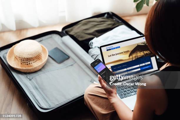 over the shoulder view of young woman booking a flight via laptop and smartphone for vacation - reserveren stockfoto's en -beelden