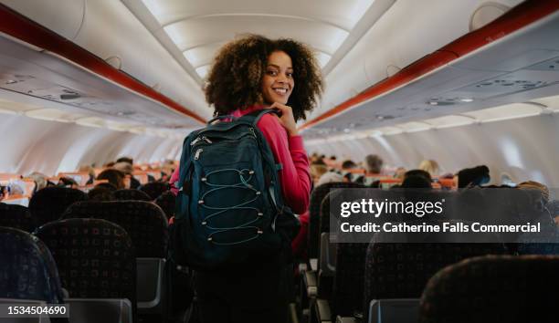 a woman boards a commercial aircraft with hand luggage. she glances over her shoulder and smiles at the camera. - production crew stock pictures, royalty-free photos & images