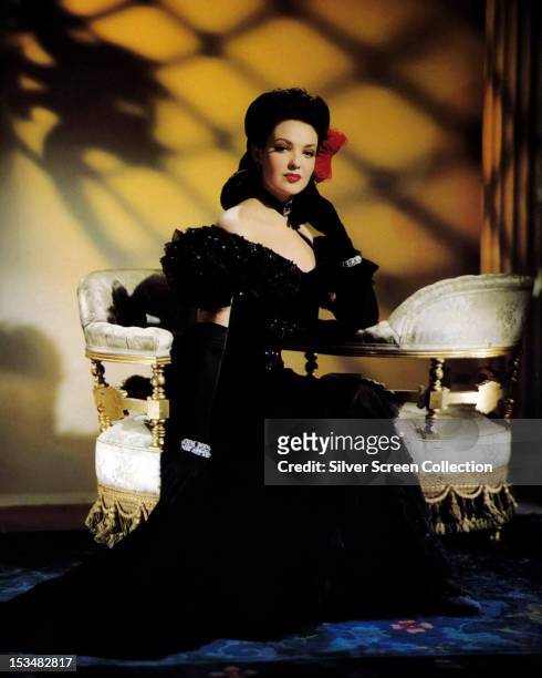 American actress Gene Tierney sitting on a love seat in a black evening gown and evening gloves, circa 1945.