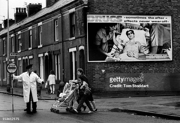 Lollipop man guides children to school across a road in Moss Side, an inner city district of Manchester. A giant poster warning of the dangers of...
