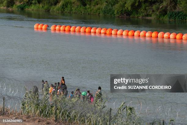 Migrants walk by a string of buoys placed on the water along the Rio Grande border with Mexico in Eagle Pass, Texas, on July 16, 2023. The buoy...