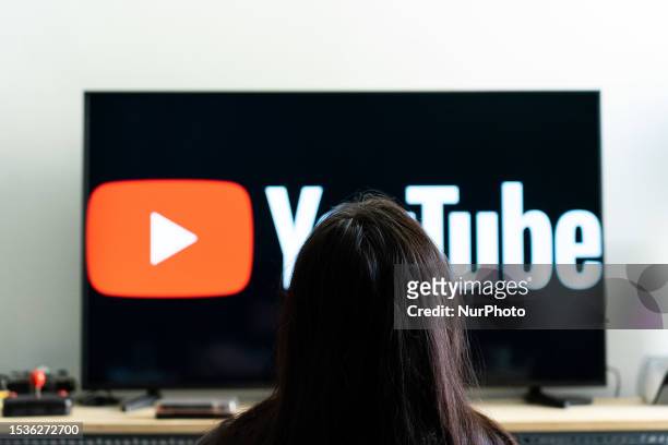 Woman seen sitting in front of a tv screen that shows the logo of You Tube platform.