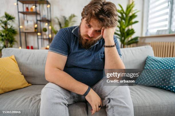 trauriger junger mann sitzt zu hause auf dem sofa - bedauern stock-fotos und bilder