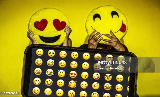 In this photo illustration, emojis are displayed on a mobile phone screen in front of a computer screen displaying a picture of people holding emoji...