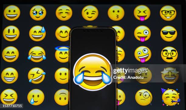 In this photo illustration, a laughing emoji is displayed on a mobile phone screen in front of a computer screen displaying a picture of emojis in...