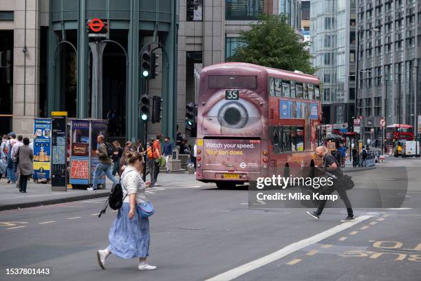 People cross Bishopsgate behind the rear of a Transport for London bus with the close up eye of a speed awareness road safety advertising campaign on...