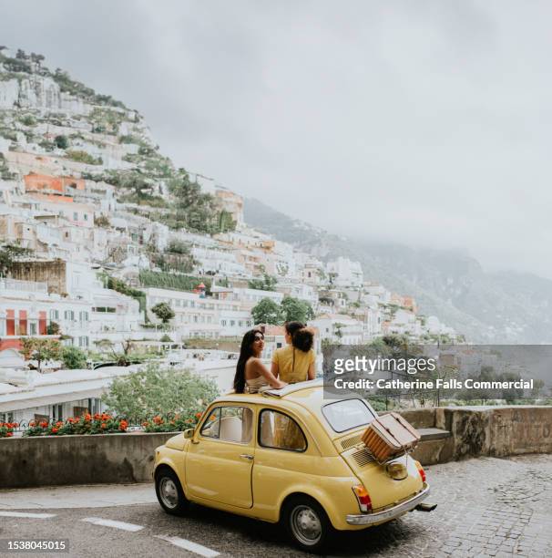 two woman stand in a vintage yellow fiat, and admire the view of positano, italy. - italie stockfoto's en -beelden