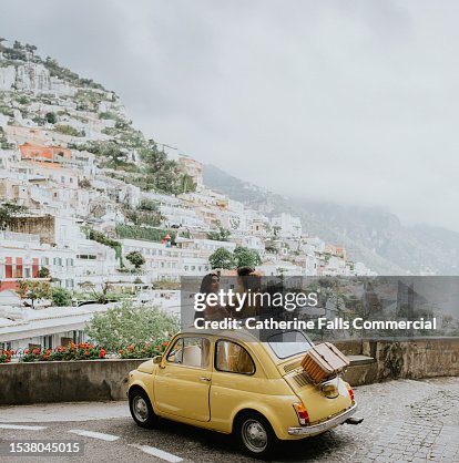 Two woman stand in a vintage yellow fiat, and admire the view of Positano, Italy.