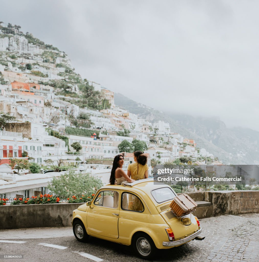 Two woman stand in a vintage yellow fiat, and admire the view of Positano, Italy.
