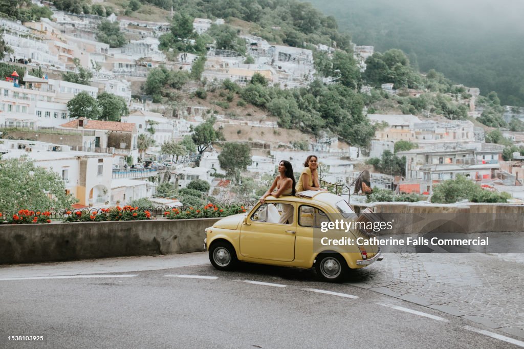 Two woman stand in a vintage yellow fiat, and admire the view of Positano, Italy.