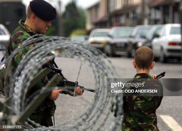 Young boy in fatigues and his replica gun sticking over his shoulder salutes a soldier when passing a check point in the Garvaghy area of Portadown...