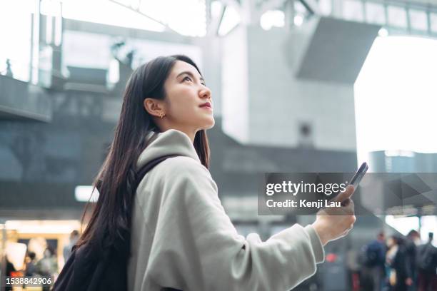 low angle of a young asian woman with backpack using smart phone in train terminal. commuting, travel and vacation concept. - japanese woman looking up stockfoto's en -beelden