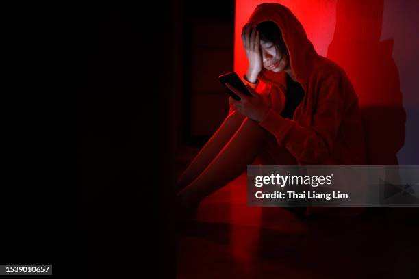 una joven asiática se sienta en el suelo, con lágrimas corriendo por su rostro mientras sostiene un teléfono inteligente. esta imagen retrata los peligros de la adicción y el ciberacoso - cyberbullying fotografías e imágenes de stock