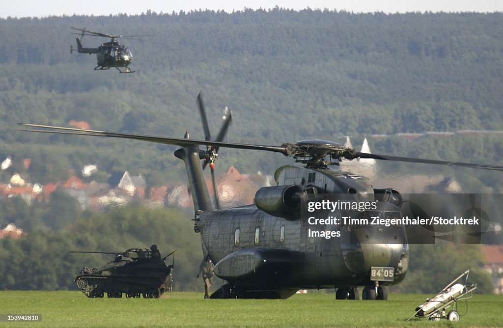 A German CH-53G helicopter unloads a Wiesel vehicle while a Bo-105 Scout helicopter watches from above, Buckeburg Airfield, Germany.