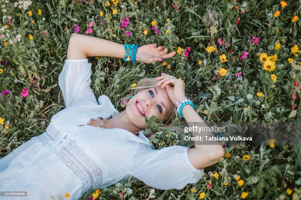 Wonderful blonde comfortable resting on field surrounded by flowers, touching forehead with hand and looking at sky with fascinating smile.