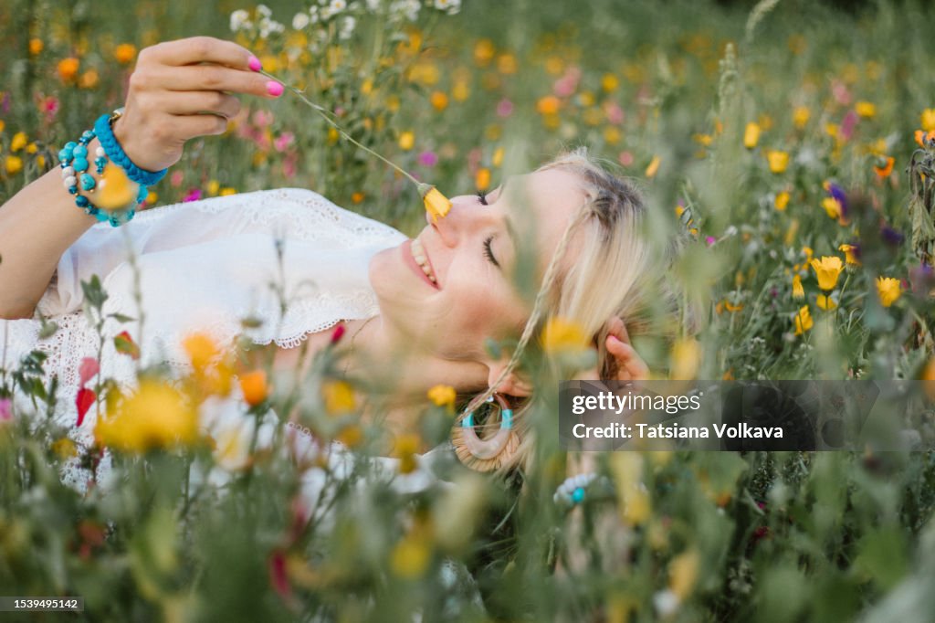 Amazing woman smelling delicate pot marigold with close eyes, lying in meadow among wildflowers and smiling.
