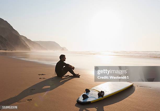 portugal, young man sitting on beach by surfboard - sitting on surfboard stockfoto's en -beelden