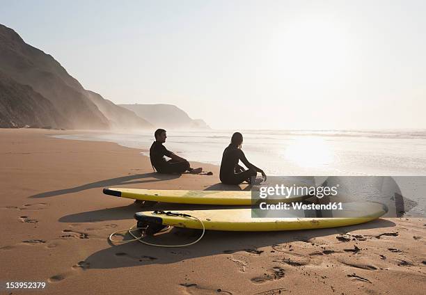 portugal, couple sitting on beach by surfboard - algarve stock-fotos und bilder