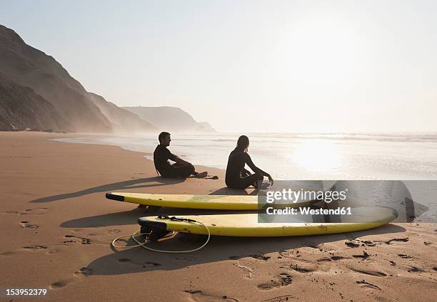 portugal, couple sitting on beach by surfboard - distrito de faro portugal fotografías e imágenes de stock