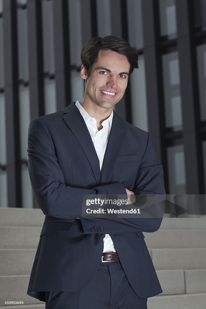 Germany, North-Rhine-Westphalica, Duesseldorf, Young businessman standing with arms crossed, smiling, portrait