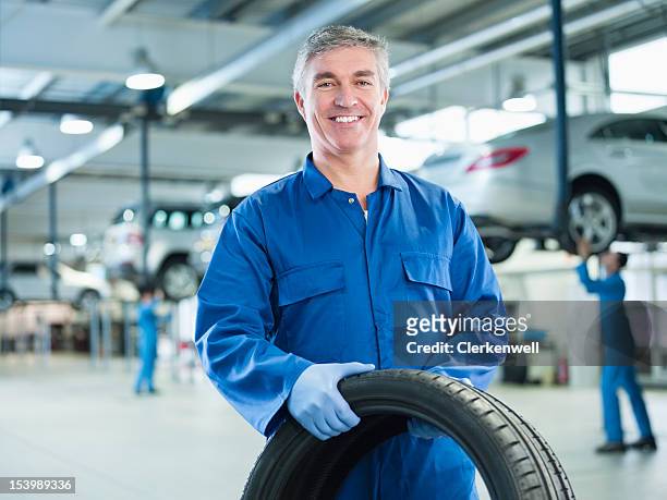 portrait of smiling mechanic holding tire in auto repair shop - 連身工作服 個照片及圖片檔