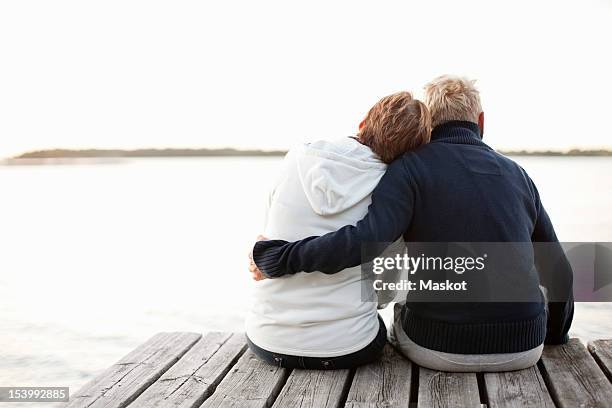 rear view of mature couple sitting on pier looking at lake - mature adult couple stockfoto's en -beelden