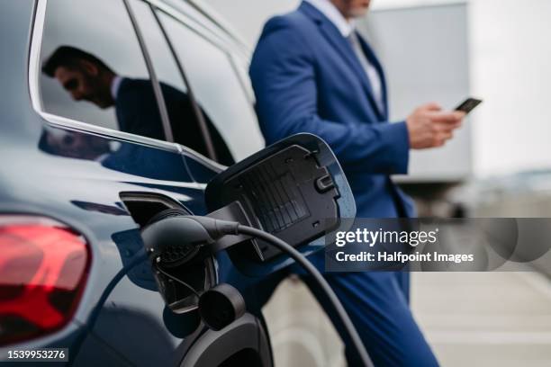 businessman calling while charging his electric car at charging station in front of his office. - hybrid car stock pictures, royalty-free photos & images