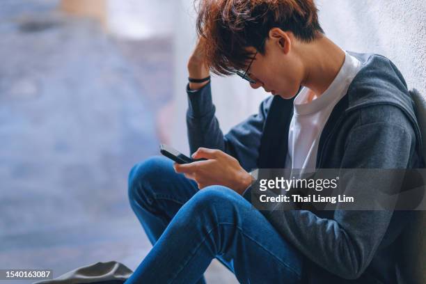 an upset asian teenage boy student, dressed in casual outfit, sits on a stairway reading on his smartphone with a backpack beside him, while dealing with the issue of cyberbullying. - teenagers only stock pictures, royalty-free photos & images
