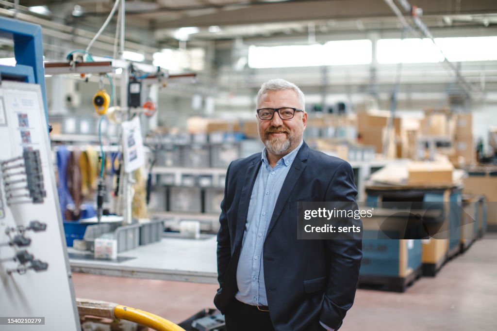 Gestão Eficiente da Fábrica: Retrato do Empresário ou CEO do Gerente Sênior Sorridente no Armazém da Fábrica Industrial