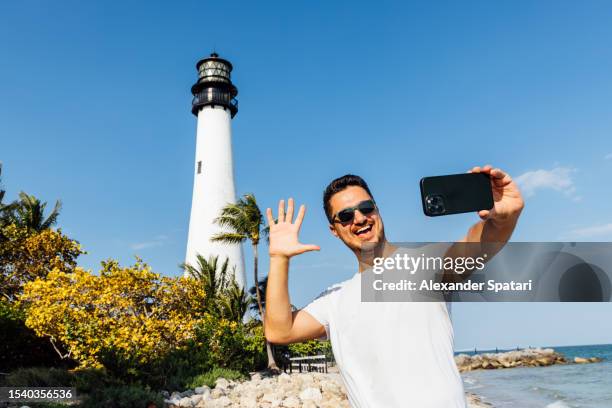 happy smiling man taking a selfie using smartphone with cape florida light lighthouse at bill baggs cape florida state park, florida, usa - florida state v miami stock pictures, royalty-free photos & images