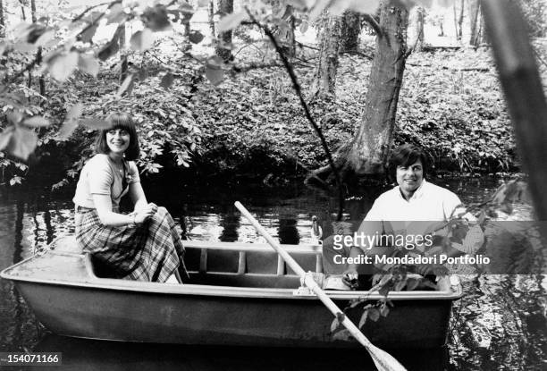 Jean Sorel and his wife Anna Maria Ferrero, both smiling, are seated on a little rowing boat along a river; the place is surrounded by a flourishing...