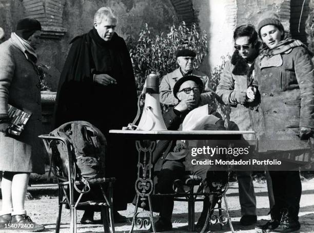 Italian director Mario Camerini seated at a table with a cigarette in his mouth and script in a hand, during a break in the filming of 'Don Camillo...