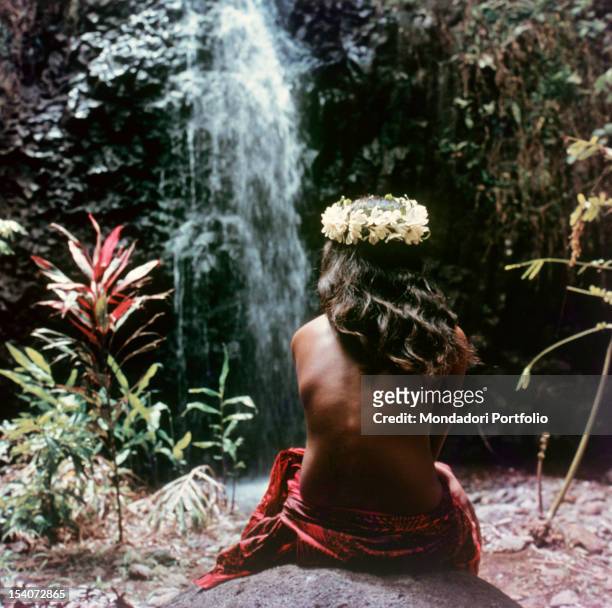 Vahine Tahitian girl wearing a crown of flowers tiare head sits on a rock and see a waterfall. Tahiti, 1964