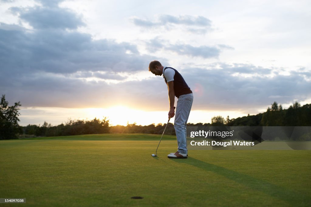 Man preparing to putt golf ball at sunset.