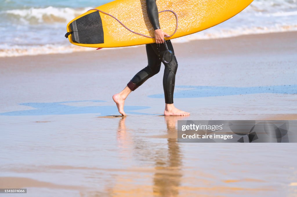 Side view of the legs of a male surfer walking on the beach sand carrying a surfboard in his hands.
