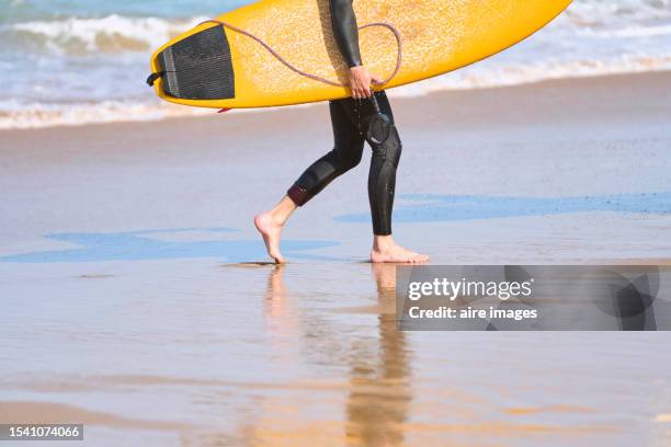 side view of the legs of a male surfer walking on the beach sand carrying a surfboard in his hands. - feet run in ocean stock pictures, royalty-free photos & images