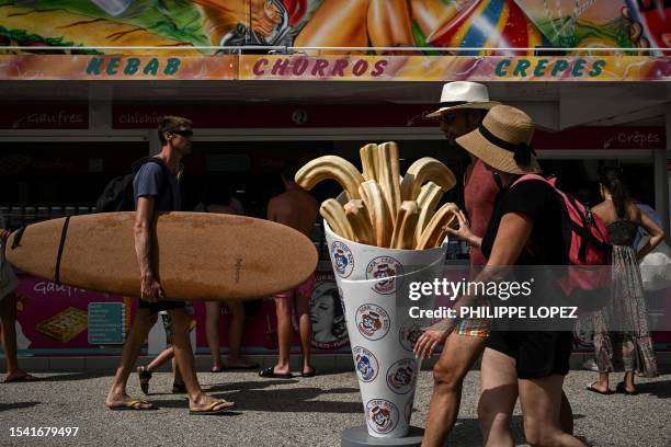 This picture taken on July 18, 2023 shows a man holding a surfboard walking past an ice cream shop near a beach in Moliets-et-Maa, south-western...