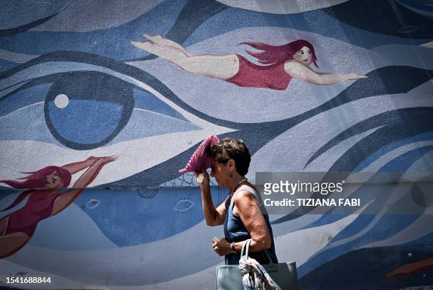 Pedestrian shelters from the sun with a handheld fan as she walks past a mural by the artists Maria Ginzburg and Gaetano Matrella, downtown Rome, on...