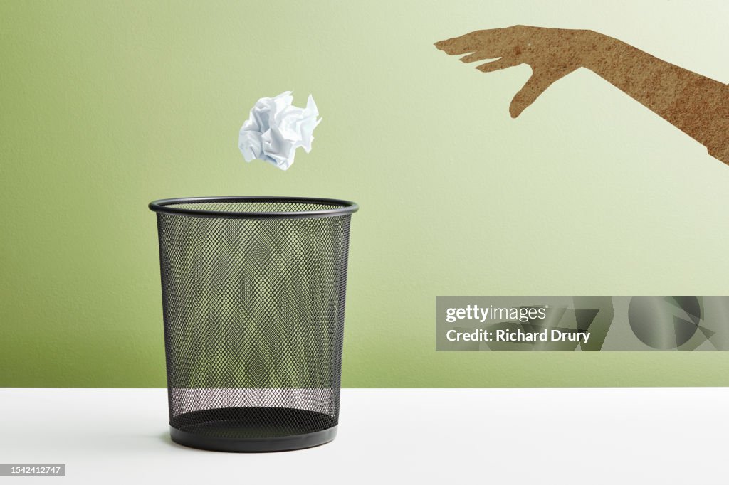 A hand throwing a crumpled paper ball into a waste paper basket