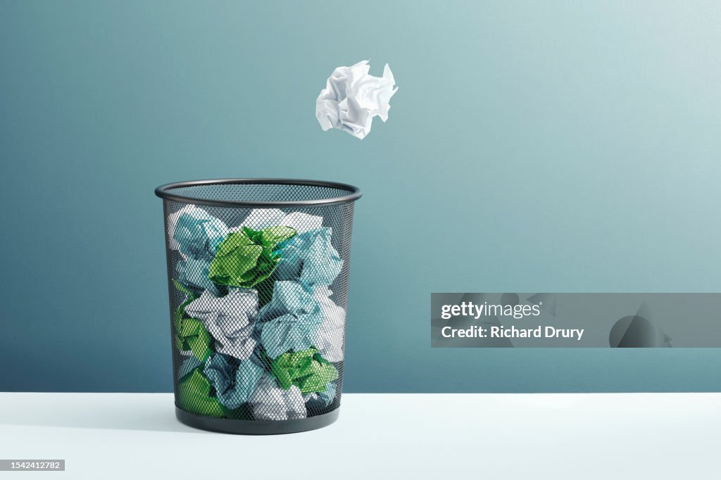 A ball of crumpled paper flying towards a full waste paper basket