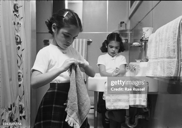 Young girl wearing a white t-shirt with the name 'Mimi' written below the crew neck, drying her hands with a towel as another young girl washes her...