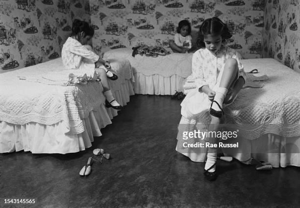 Two young girls sit on their beds as they put their shoes on, their younger sister sitting on a third bed in the background in their bedroom, United...