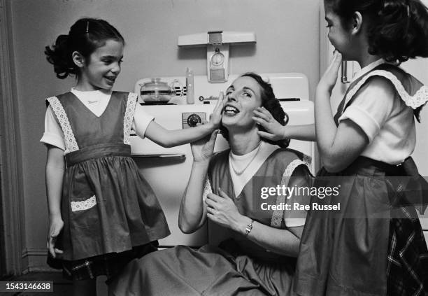 Two young girls, one with the name 'Mimi' written below the crew neck of her t-shirt, both reaching out to touch the face of their mother who sits...